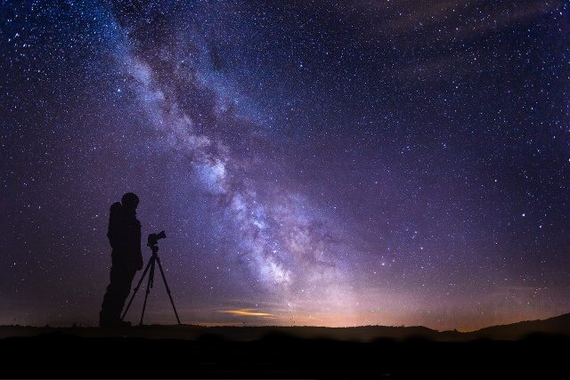 Image of a scenery showing the Milky Way galaxy arm and a silhouette of a human and a camera on tripod.